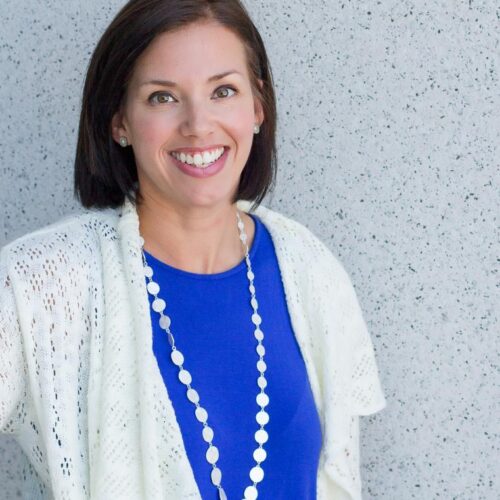 A headshot of a woman with short brown hair wearing a blue top with a white sweater