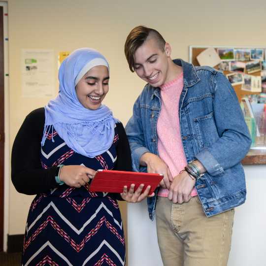 Two students stand in an office while looking at a tablet together