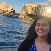 A woman smiles standing in front of a body of water with the Sydney Opera House in the background