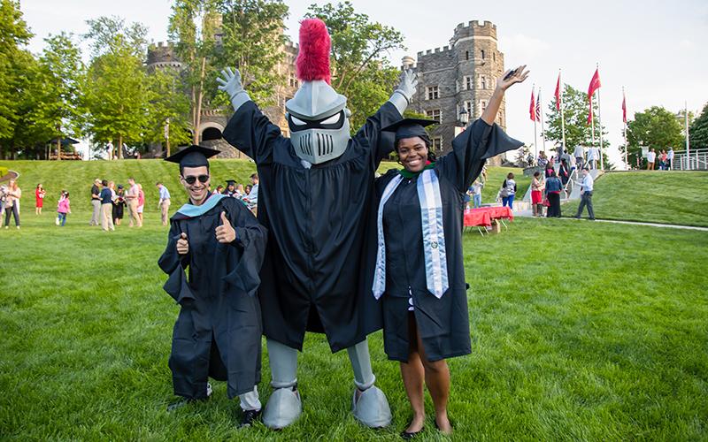 Two students in graduation caps and gowns stands with Arcadia knight mascot