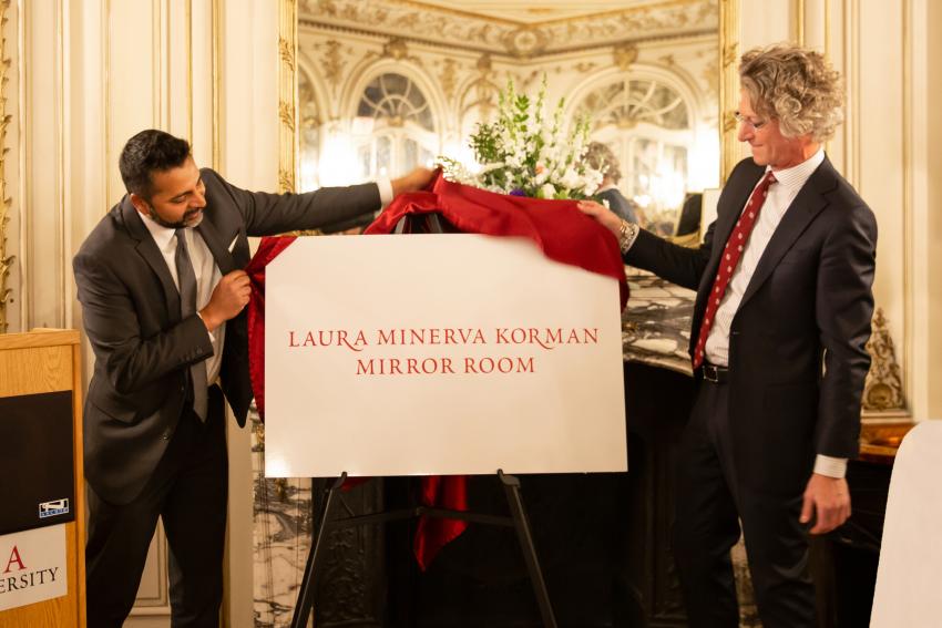 The president of Arcadia University and another man in a suit unveil a sign that says "Laura Minerva Korman Mirror Room" while standing in a room of mirrors