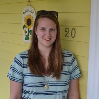 A woman in a turquoise striped shirt smiles in front of a yellow house with the number 20 on it