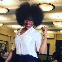 A woman with an afro smiles at the camera wearing eyeglasses and a white short-sleeved blouse