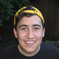 A headshot of a person with brown curly hair, a backwards yellow baseball cap, and a black t-shirt