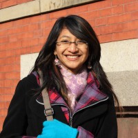 A young woman smiles at the camera wearing glasses, a purple scarf, a black coat with a red and purple inner collar, and blue gloves