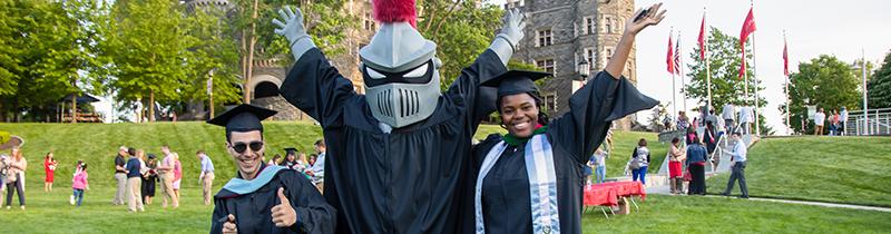 Two students in graduation caps and gowns stands with Arcadia knight mascot