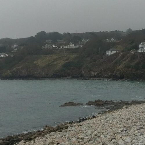 A landscape of a beach that is rocky in the foreground and hilly with sporadic homes in the background