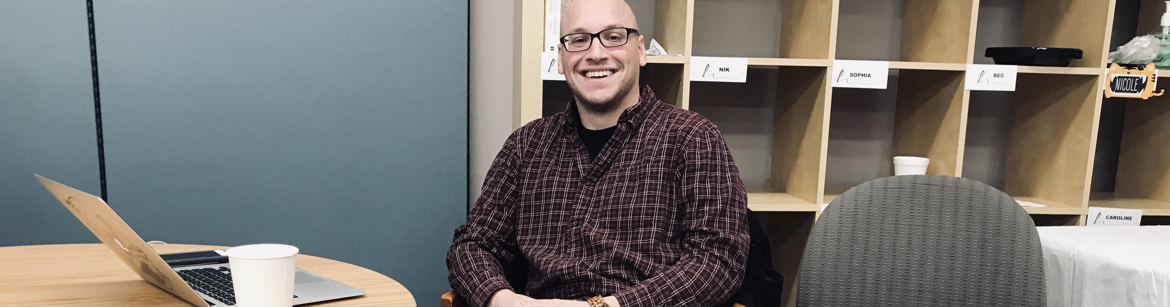 A man wearing glasses and a purple plaid shirt smiles at the camera with a laptop and white cup on the table beside him and cubbies behind him