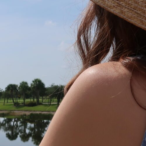 A woman's back is turned to the camera as she gazes at a body of water reflecting the trees beyond