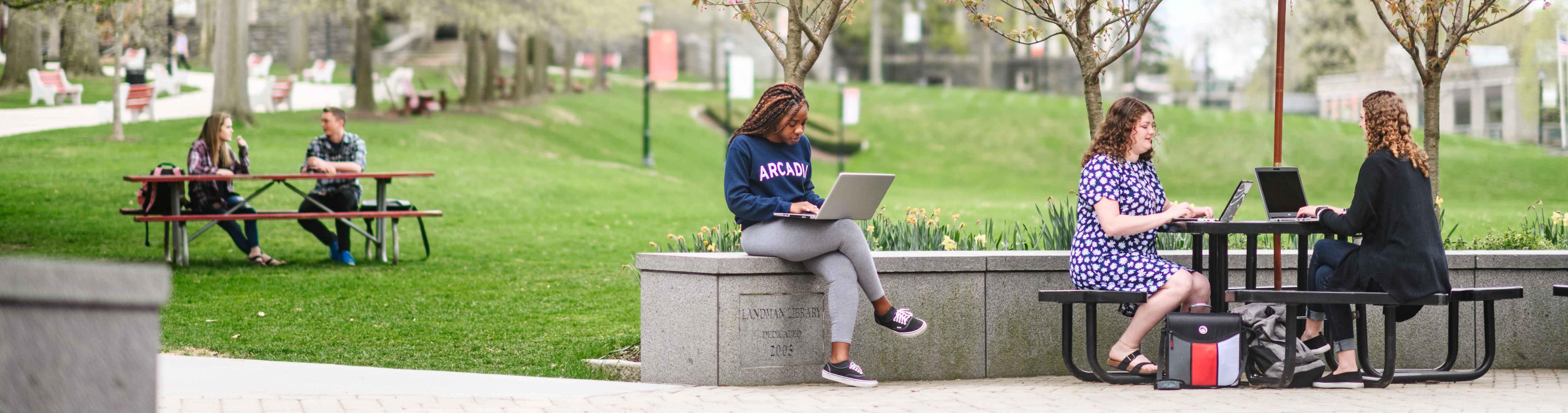 Photo of students sitting outside on campus talking, studying, and on their computers.