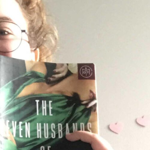 A person holding a book called "The Even Husbands" in front of their head