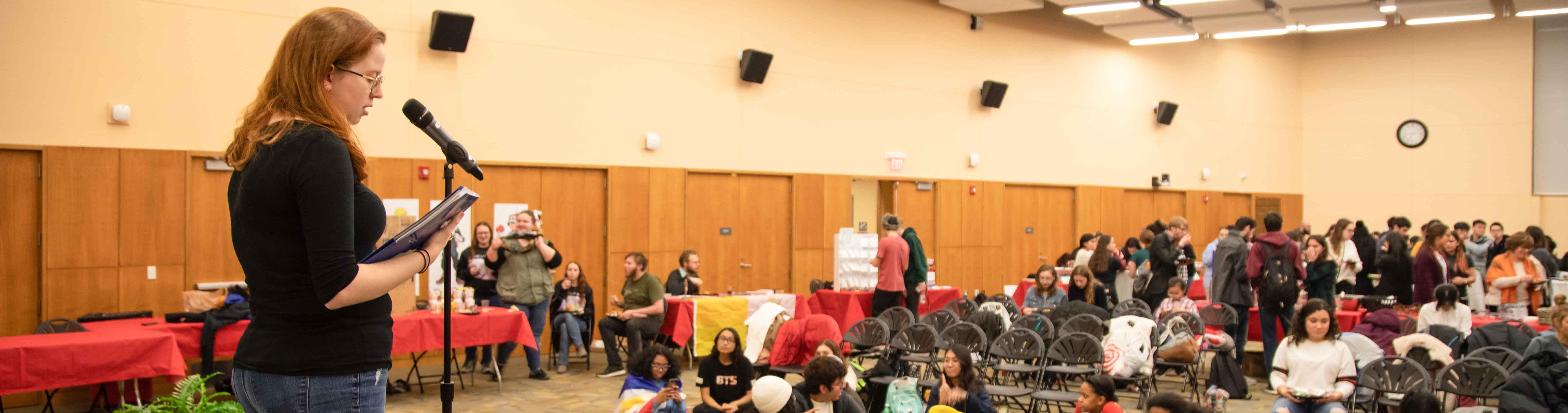 A woman speaking at a microphone in the Great Room to students and faculty.