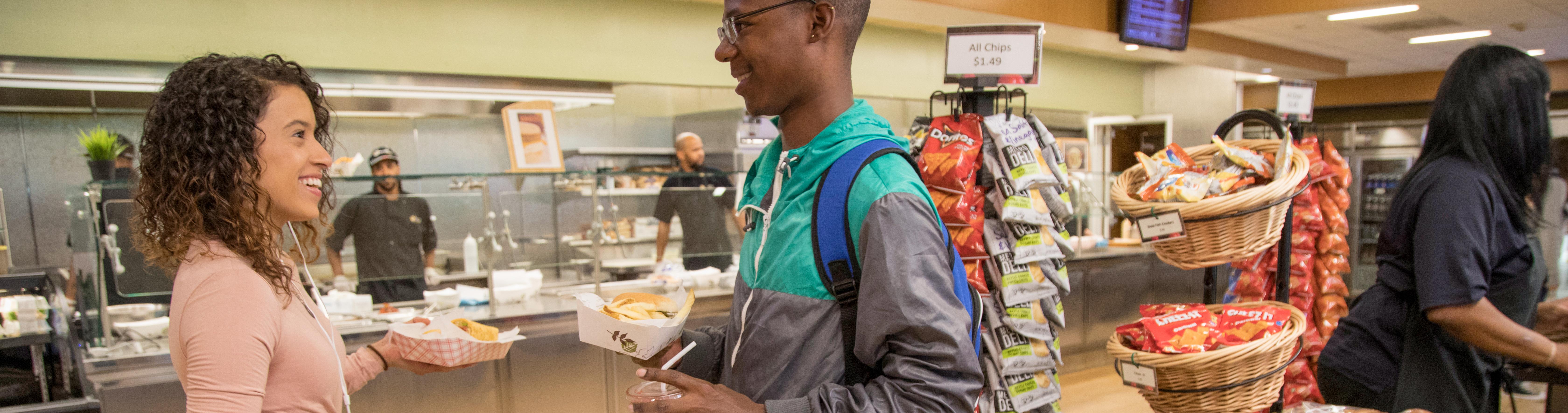 Two students smile at each other while holding food, a food counter behind them