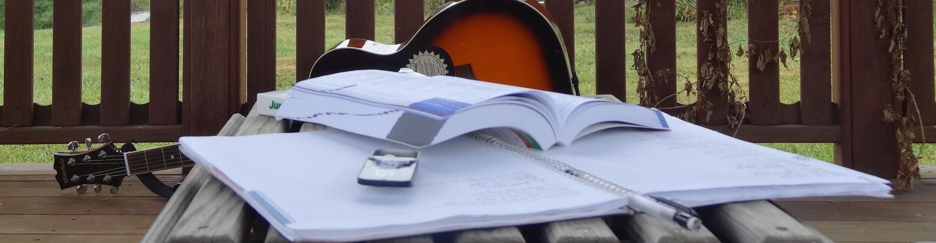 A guitar is propped on its side on a porch with an open book and notebook in front of it.
