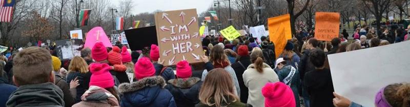 Sign that says "I'm with Her" pointing towards the crowd surrounding it in a street.