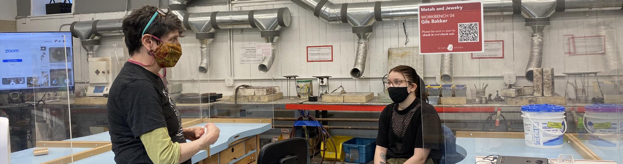A masked teacher and student sit in the metal work lab.