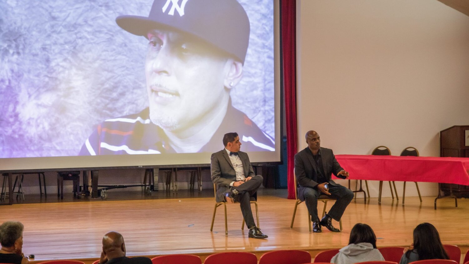 Two men sit on stage and give a speech with a video playing behind them