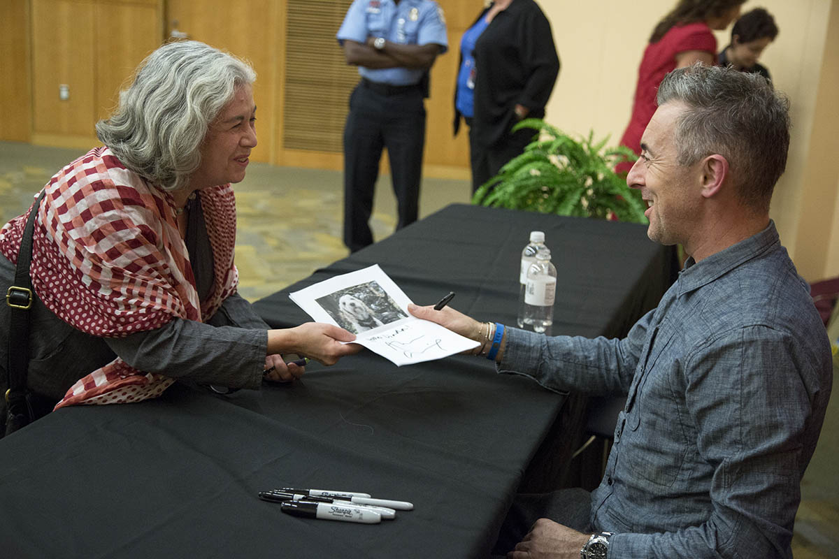 Alan Cumming signing and greeting a person who has a paper in their hand with an image of dog
