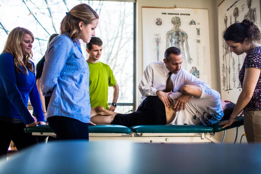 Physical therapy students stand around an examination table as an professor teaches.