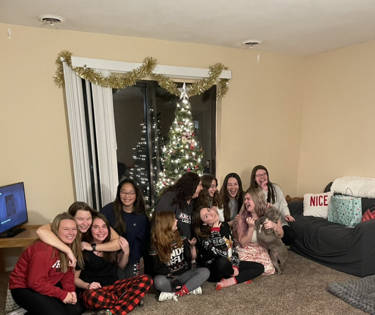 A group of students wearing pajamas, laughing while seated on the floor, a Christmas tree behind them