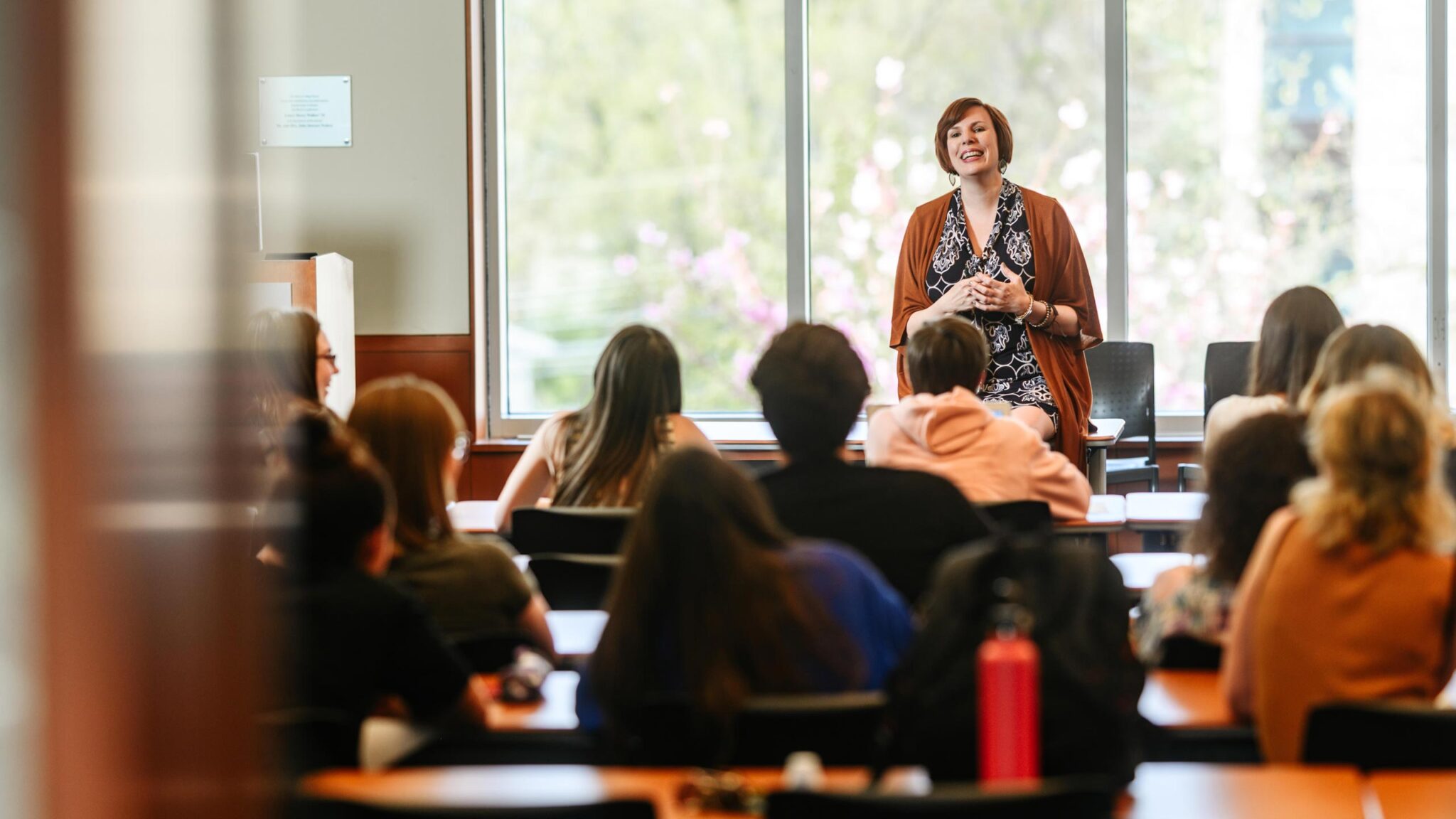 A professor smiles while teaching in the front of a classroom full of students.