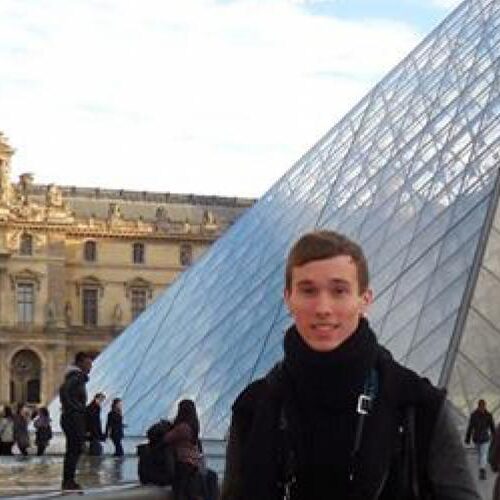 A student in a black jacket stands in front of the Louvre museum in Paris, which has a pyramid structure