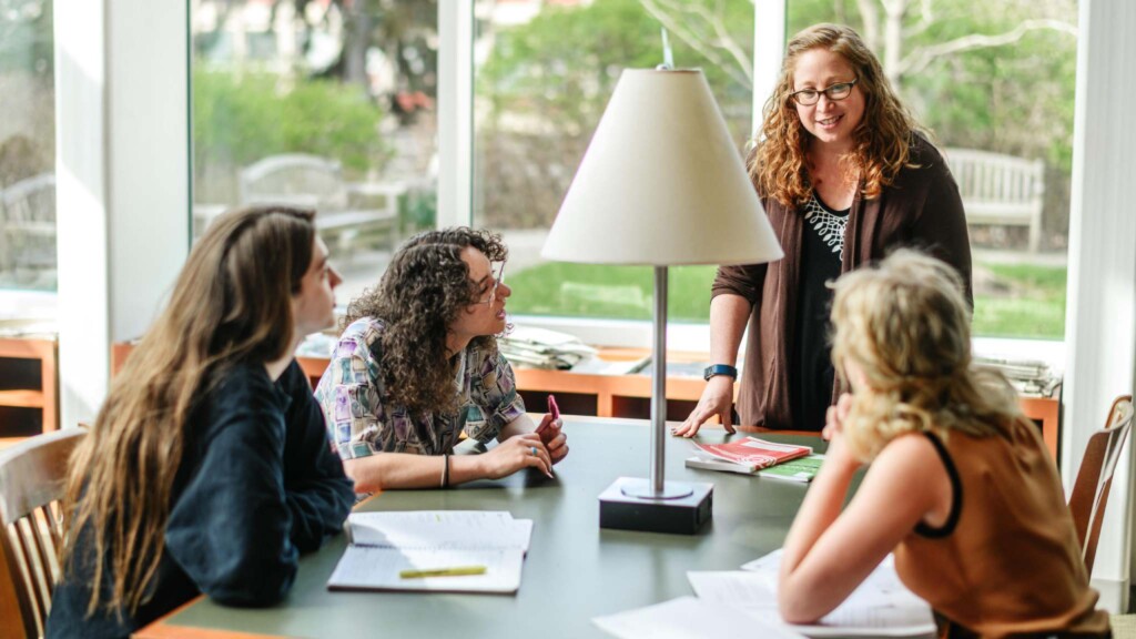 English professor and students discuss a lesson around a table in front of a sunny window.