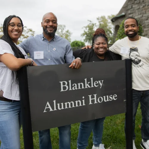 Alumni smiling at the camera and standing with the "Blankley Alumni House" sign.