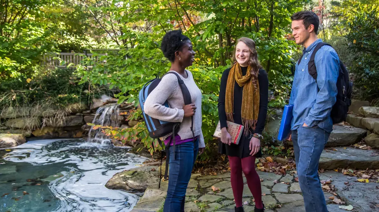 Smiling students stand next to Easton Pond wearing bookbags and carrying books