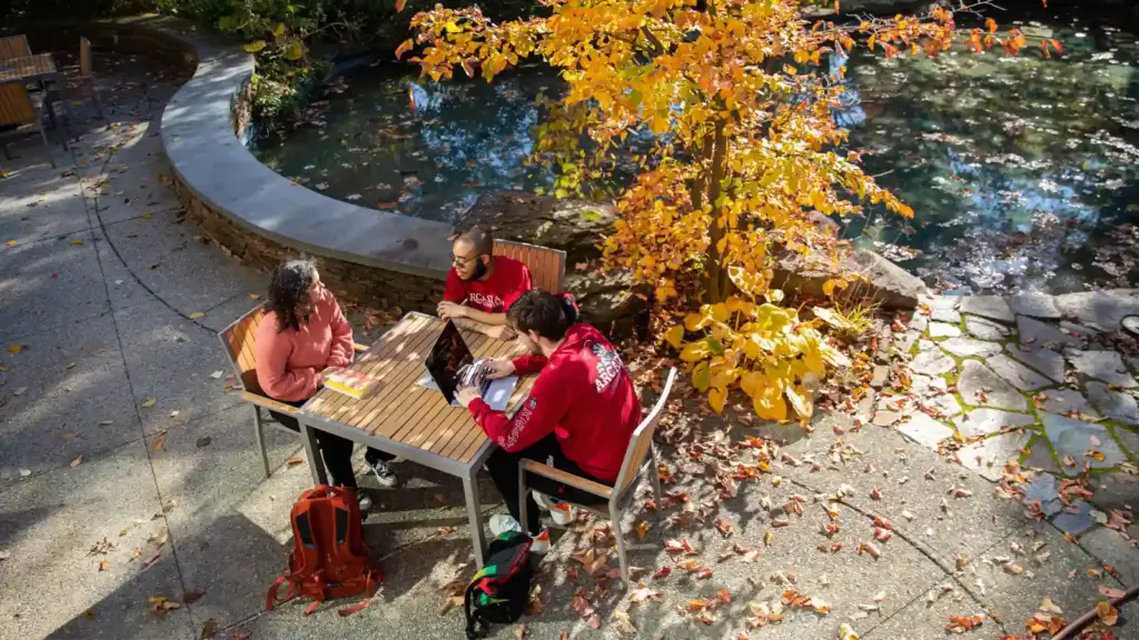 Students talk and do work while seated at a table outside.