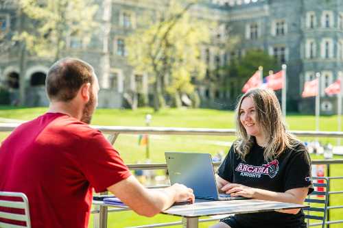 Two students sit opposite each other working on a laptop at an outdoor table in the commons