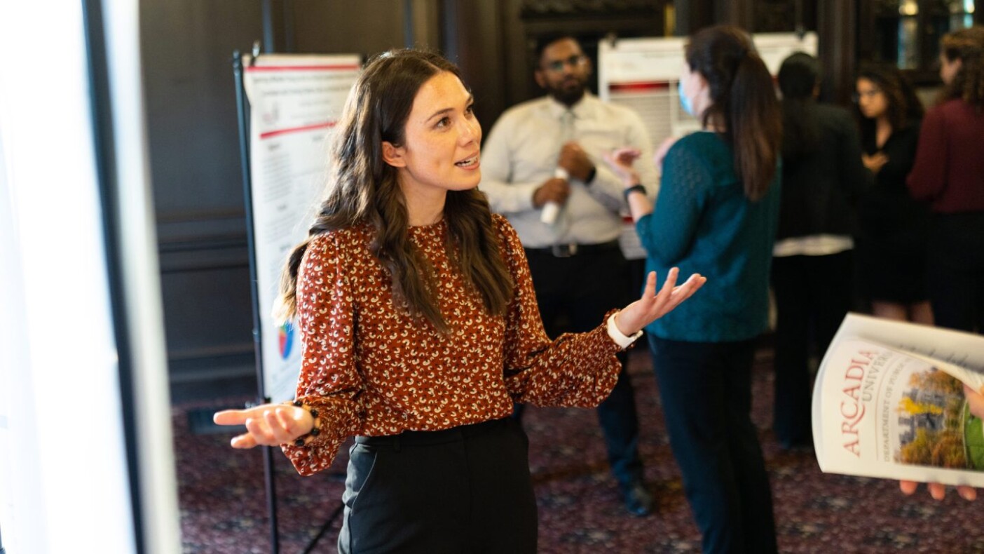 A woman in business attire speaks at an event hall filled with chart displays.