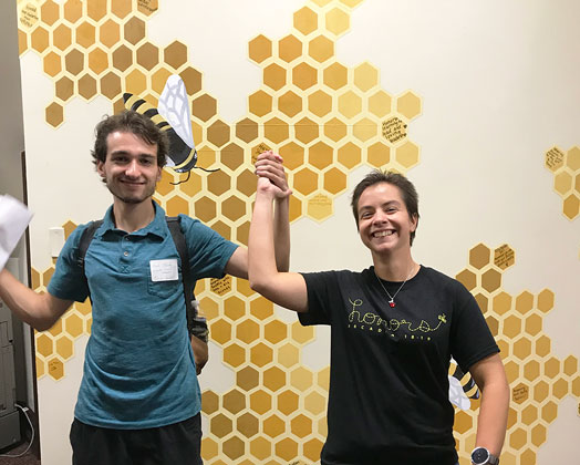 Two Honors students stand by the bee wall at the Hive.d in front of the bee