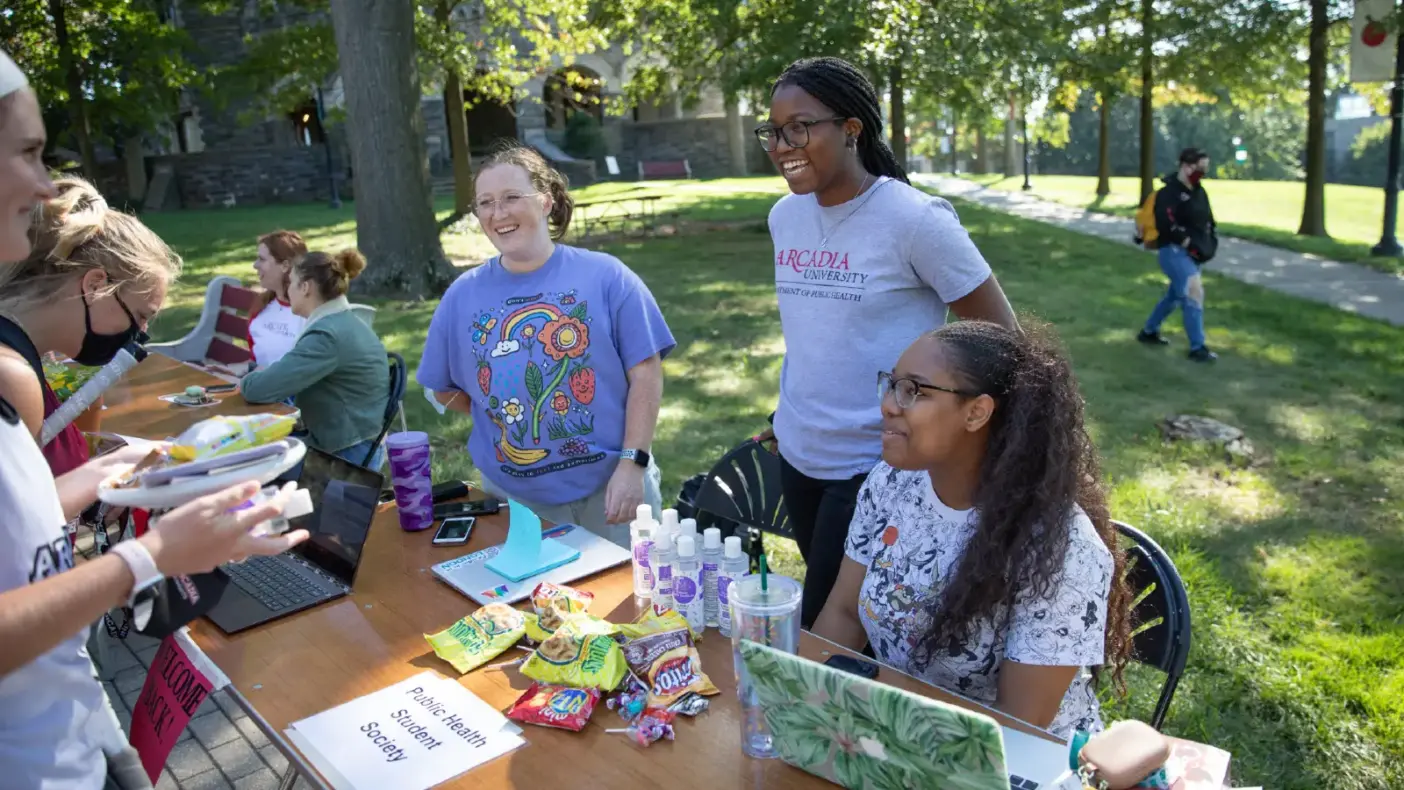 Three students sit behind table and talk to other students about Public Health Student Society at the Activities fair.