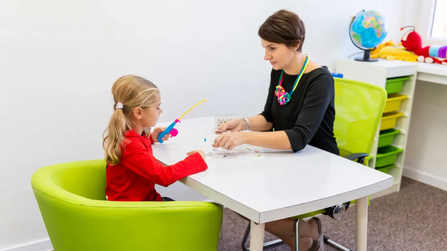 A woman helping a child with motor skills by having her use a toy fishing rod to pick papers up