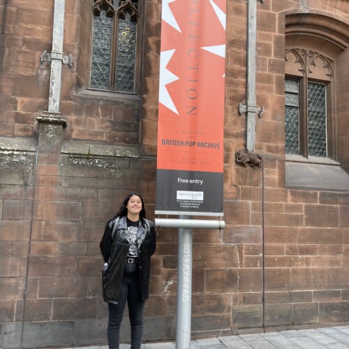 Young woman dressed all in black stands next to a sign for the British Pop Archive at the John Rylands Research Library in Manchester, England.