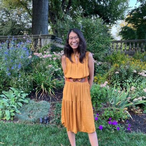 A female student with long dark hair stands in a garden smiling wearing a golden dress with a brown belt