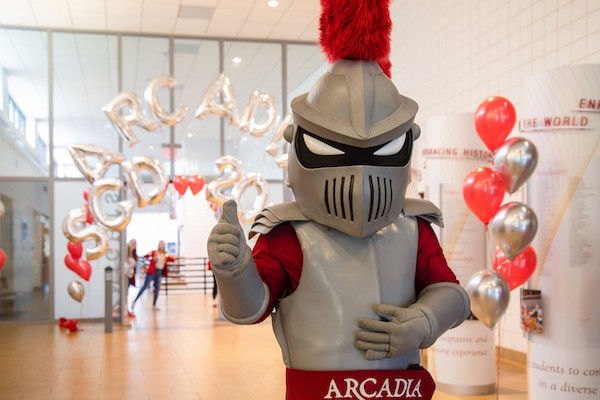 Arcadia Knight mascot gives a thumbs up in a sunny hall filled with balloons