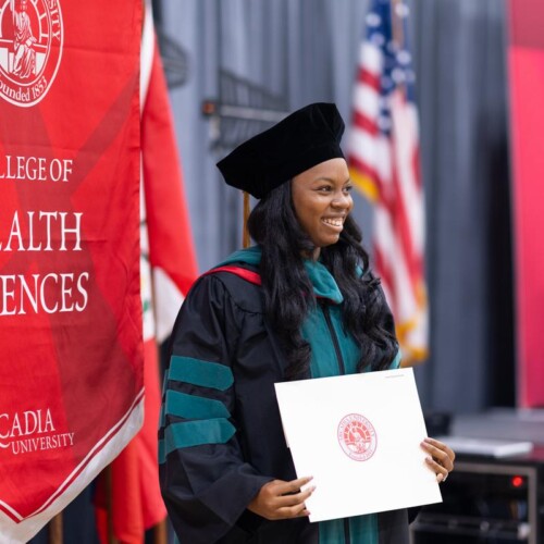A graduate holds the diploma in front of the Health Sciences banner at DPT commencement