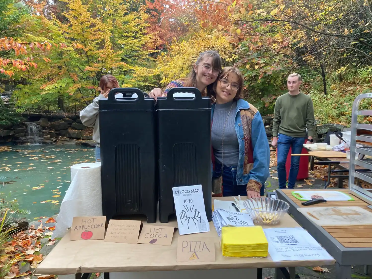 Two students smiling at hot cider, cocoa, and pizza stand.