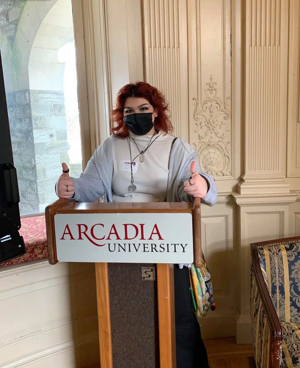 A student wearing a face mask stands at an Arcadia University podium and gives two thumbs up