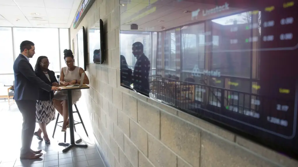 Three students gathered around a laptop next to T V with stock exchange information displayed.