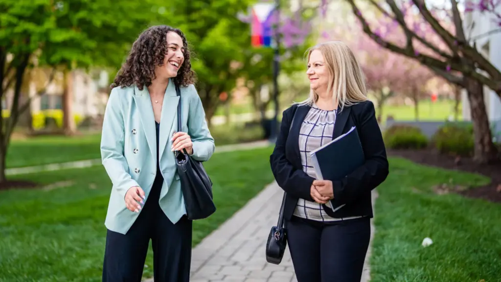 Two students chat and laugh as they walk through campus on a cloudy summer day.