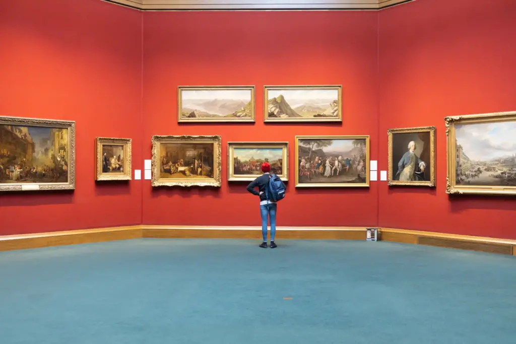 A student with a filled backpack stands in front a gallery wall of classical European art gilded in golden frames.