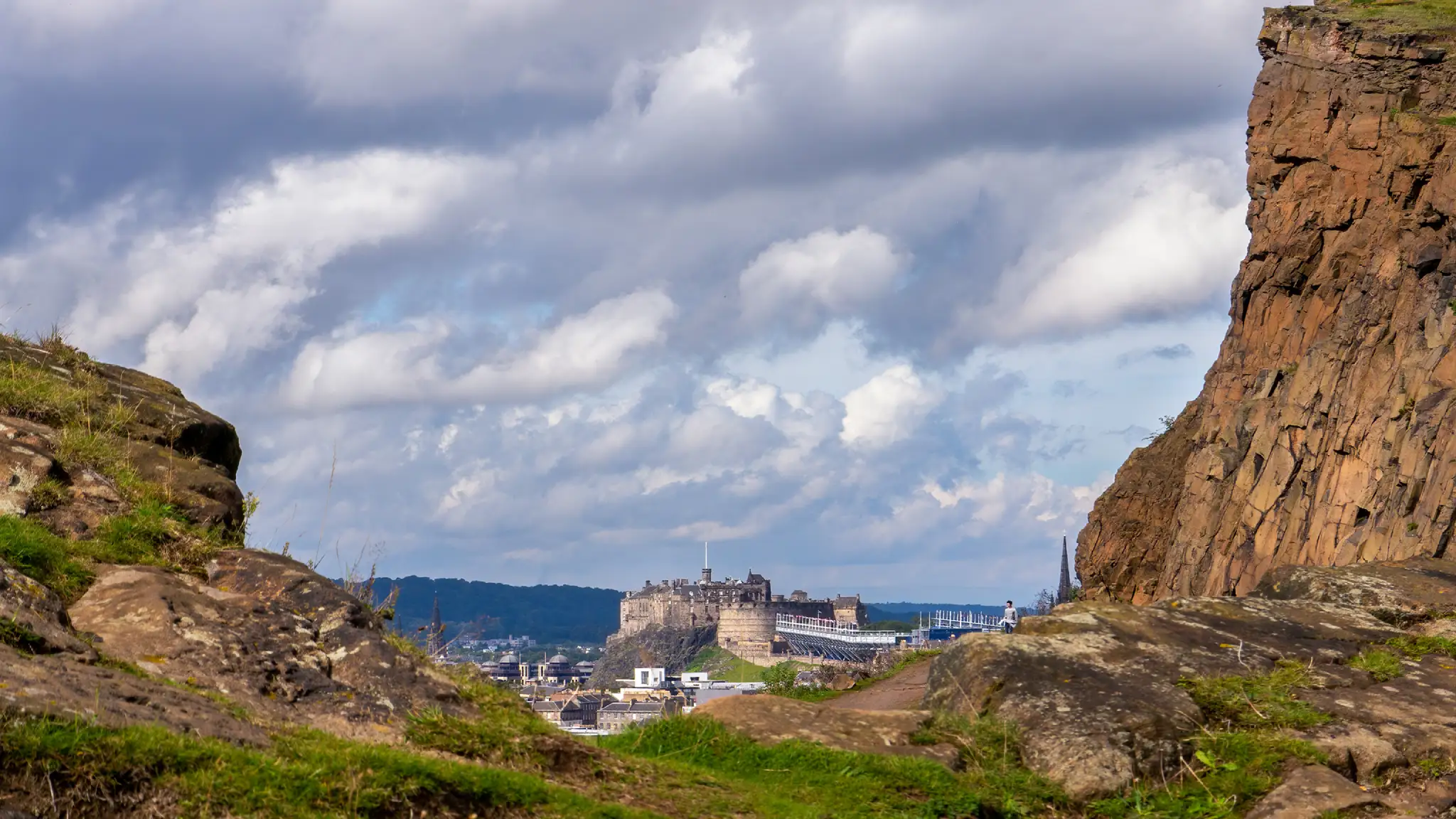 A dramatic view of a castle, mountains, and clouds around Arthur's Seat in Edinburgh, Scotland.