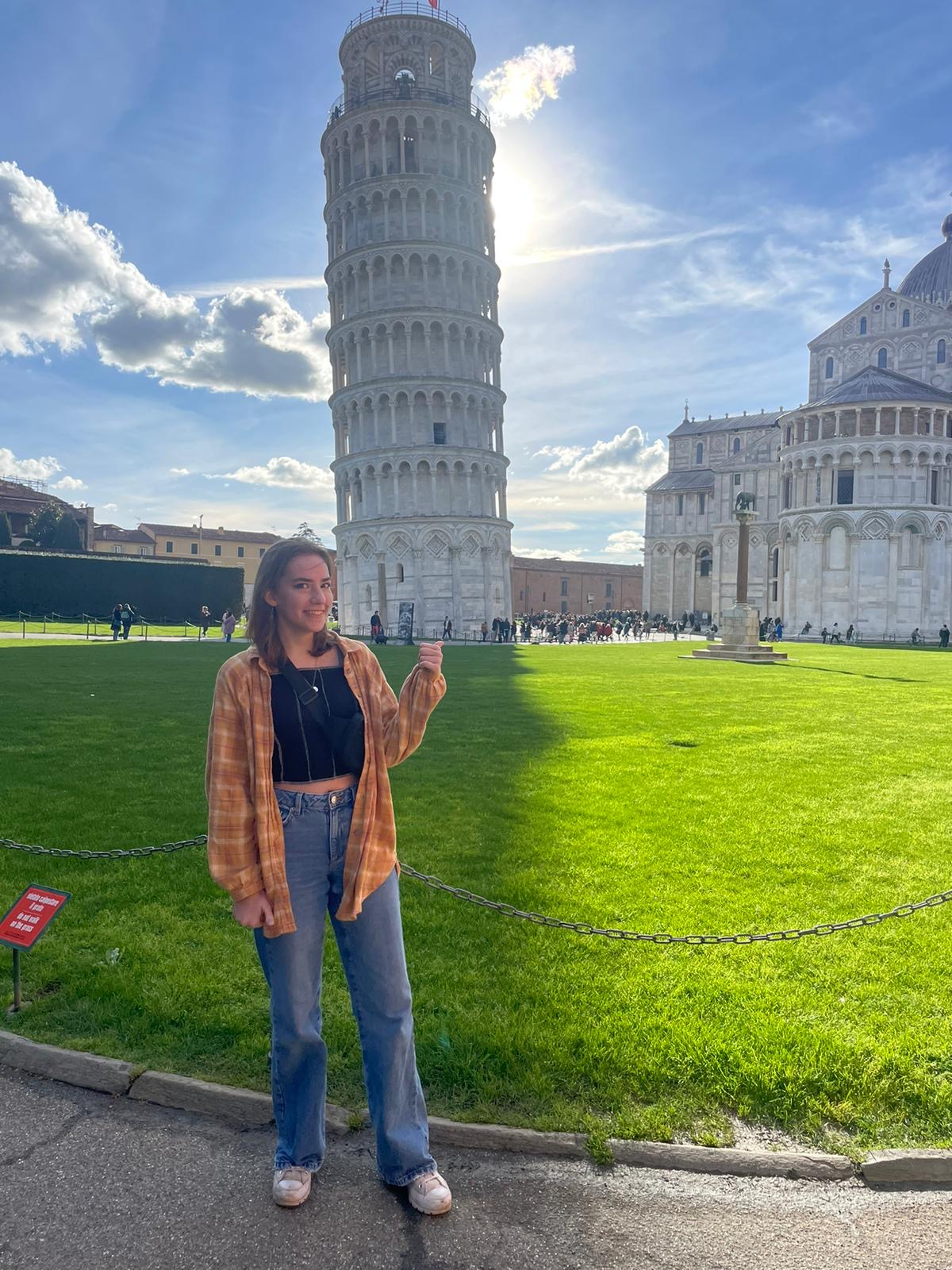 Student smiling and standing in front of the Leaning Tower of Pisa, wearing jeans and an orange flannel