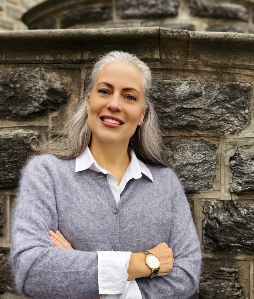 woman with silver and white hair smiling with her arms crossed in front of a stone wall