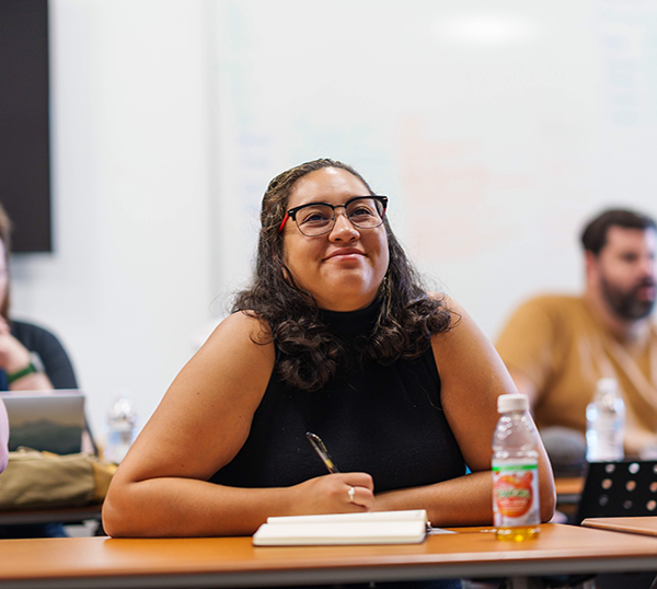An undergraduate student sits at a classroom table during a lecture and smiles.