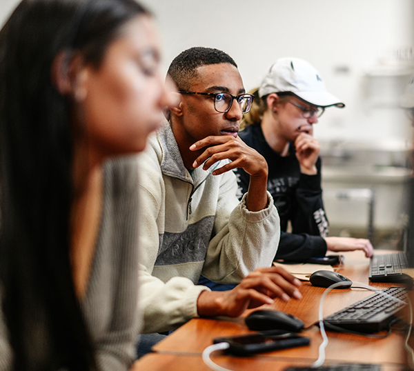 Computer science students sit in a row and lean into monitors as they work in class.