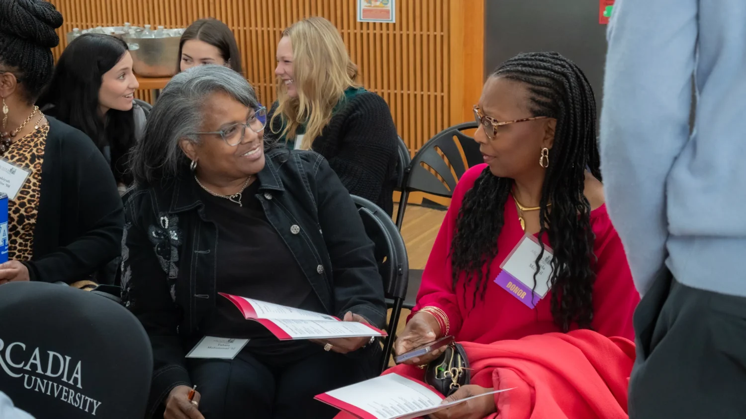 Two Black women sitting in black chairs facing each other and talking.